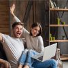 young smiling couple sitting on couch at home in casual outfit, love and romance, woman and man embracing, wearing jeans, spending relaxing time together, holding laptop, happy emotional