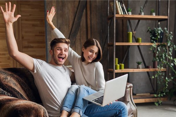 young smiling couple sitting on couch at home in casual outfit, love and romance, woman and man embracing, wearing jeans, spending relaxing time together, holding laptop, happy emotional