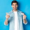 Amazed handsome man shopping online, holding mobile phone and credit card, smiling while paying for internet purchase, standing over blue background