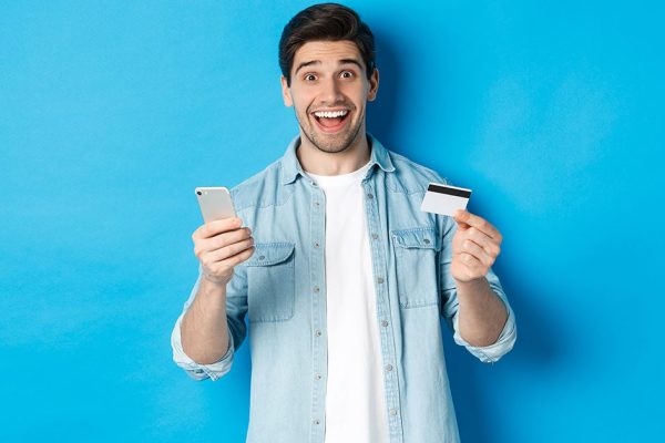 Amazed handsome man shopping online, holding mobile phone and credit card, smiling while paying for internet purchase, standing over blue background