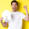 Real estate concept. Cheerful man showing paper house model and making fist pump, paid mortgage, yellow background