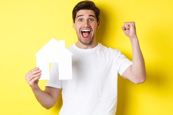 Real estate concept. Cheerful man showing paper house model and making fist pump, paid mortgage, yellow background