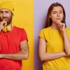 Horizontal shot of pensive millennial couple hold chins and look with displeasure at camera, wear bright casual clothes, stand together shoulder to shoulder against purple and yellow background