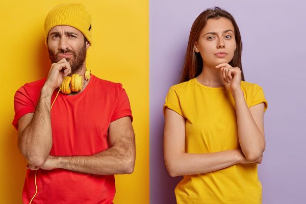 Horizontal shot of pensive millennial couple hold chins and look with displeasure at camera, wear bright casual clothes, stand together shoulder to shoulder against purple and yellow background