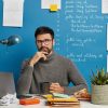 Pensive bearded employee in casual turtleneck, thinks on corporate information, holds mug of tea, poses in coworking space, sits in front of laptop computer against blue background. Software developer