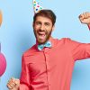 Photo of joyful birthday guy dances on party, enjoys loud music wears party cap elegant pink shirt with bowtie poses against blue wall with colorful balloons on left side. Lets have fun. Entertainment