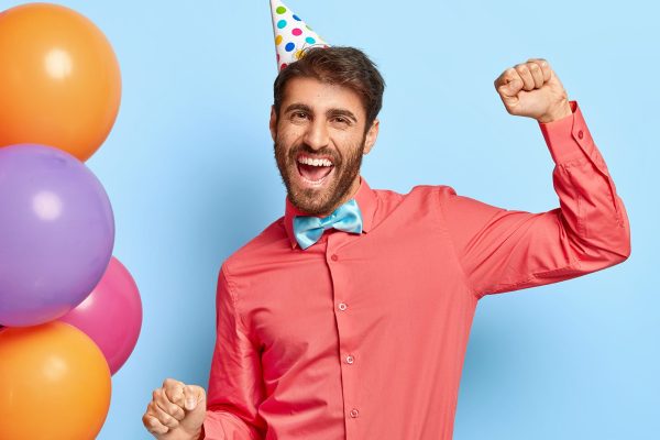 Photo of joyful birthday guy dances on party, enjoys loud music wears party cap elegant pink shirt with bowtie poses against blue wall with colorful balloons on left side. Lets have fun. Entertainment