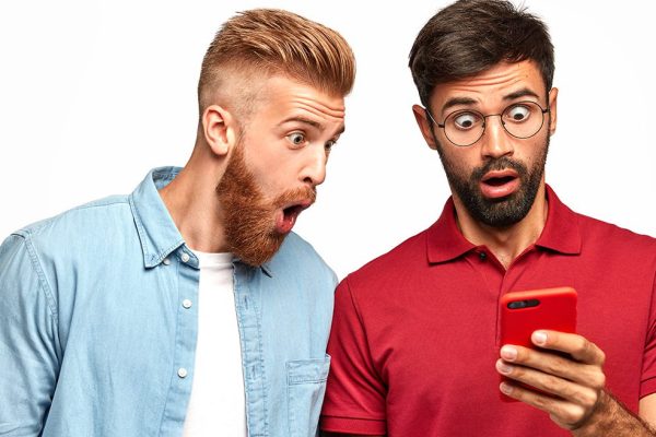 Photo of amazed surprised unshaven men stare at screen of smart phone, read sudden news on website, connected to high speed internet. Ginger bearded man and his brother isolated over white wall