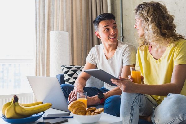 young couple sitting on sofa at home working online on laptop and tablet, using internet, smiling, happy together, having fun, man and woman in love, family leisure time, having breakfast in morning