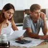 Portrait of young couple: female reading attentively document and male sitting in front of open laptop and chatting with business partner over smart phone, being busy with making financial report