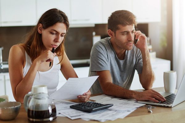 Portrait of young couple: female reading attentively document and male sitting in front of open laptop and chatting with business partner over smart phone, being busy with making financial report