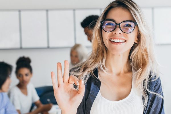 Close-up portrait of pretty female manager from sales department. Indoor photo of smiling girl working in office with discussing people on background..