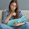 Studying online concept. Serious young woman being busy with remote freelance project, sits at comfortable sofa, writes notes, holds textbook, use laptop computer at home with wireless internet