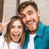 Smiling beautiful girl and her handsome boyfriend in casual summer clothes. Happy family taking selfie self portrait of themselves on smartphone camera. Shows peace sign and winking in the street