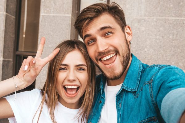 Smiling beautiful girl and her handsome boyfriend in casual summer clothes. Happy family taking selfie self portrait of themselves on smartphone camera. Shows peace sign and winking in the street