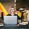 funny happy excited young pretty woman sitting at table in black shirt working on laptop in co-working office, wearing glasses