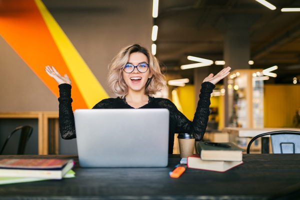 funny happy excited young pretty woman sitting at table in black shirt working on laptop in co-working office, wearing glasses