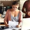 Young pretty woman wearing glasses on her head smiling happily while reading document saying that bank approved their mortgage application, her husband standing on background and drinking coffee