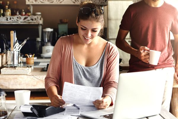 Young pretty woman wearing glasses on her head smiling happily while reading document saying that bank approved their mortgage application, her husband standing on background and drinking coffee